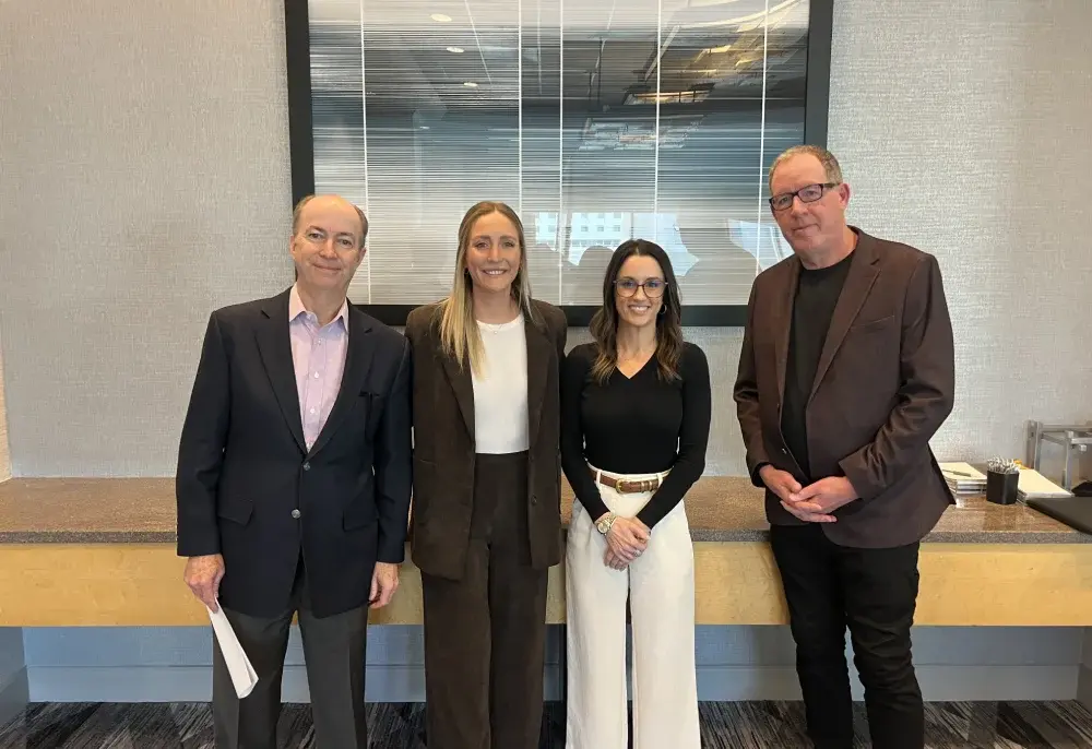 Four individuals stand in front of a stylish backdrop, dressed in formal attire, with a modern decor visible in the background.