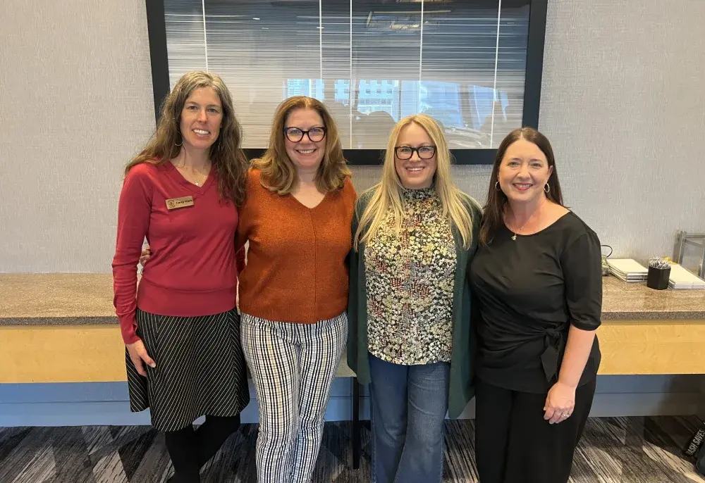 Four women stand together smiling in a modern hotel lobby, dressed in colorful outfits, with a welcoming atmosphere.