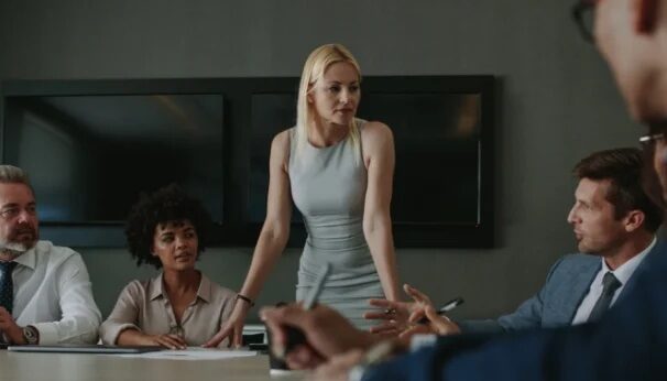 A diverse group of professionals meeting around a table with one standing professional.