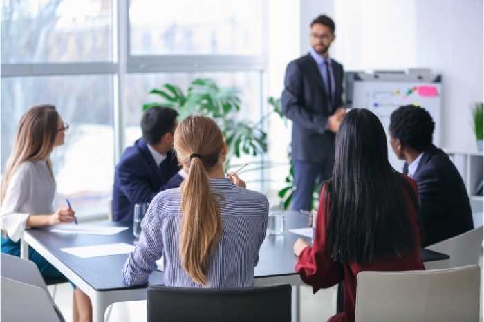 A diverse group of professionals meeting around a table where one standing human engages using visuals.